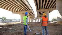 Männer in Sicherheitswesten, vermutlich Ingenieure, stehen unter dem Rohbau einer Brücke